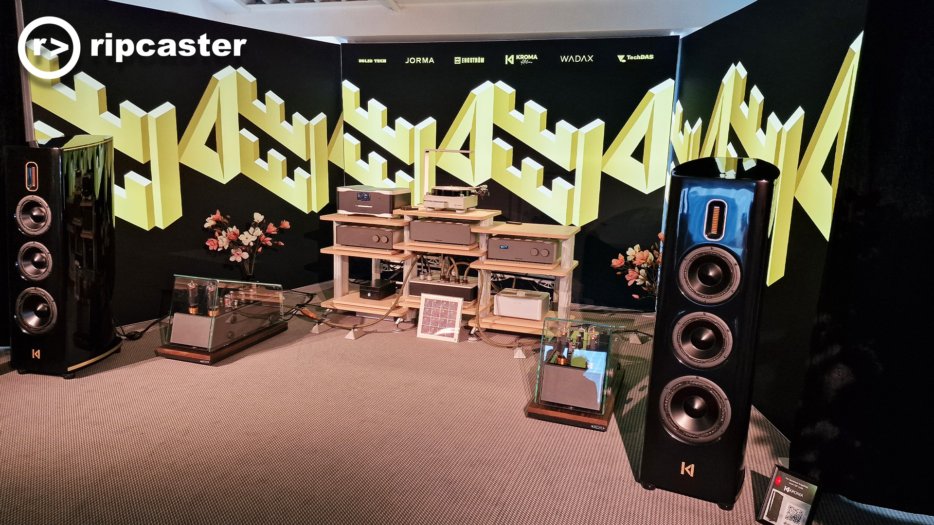 Various bits of HiFi kit on racks between two black floorstanding speakers with a striking backdrop of geometric letters
