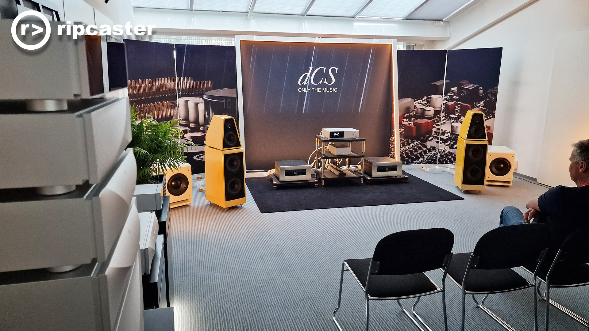 Yellow speakers either side of a range of HiFi kit.  Three chairs are viewed from behind in the foreground.  one has a man sitting in it.