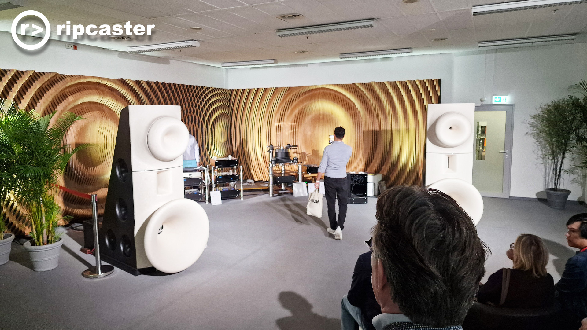White funnel speakers in a listening room with a lot of other HiFi kit and people in the foreground with one man taking a photo