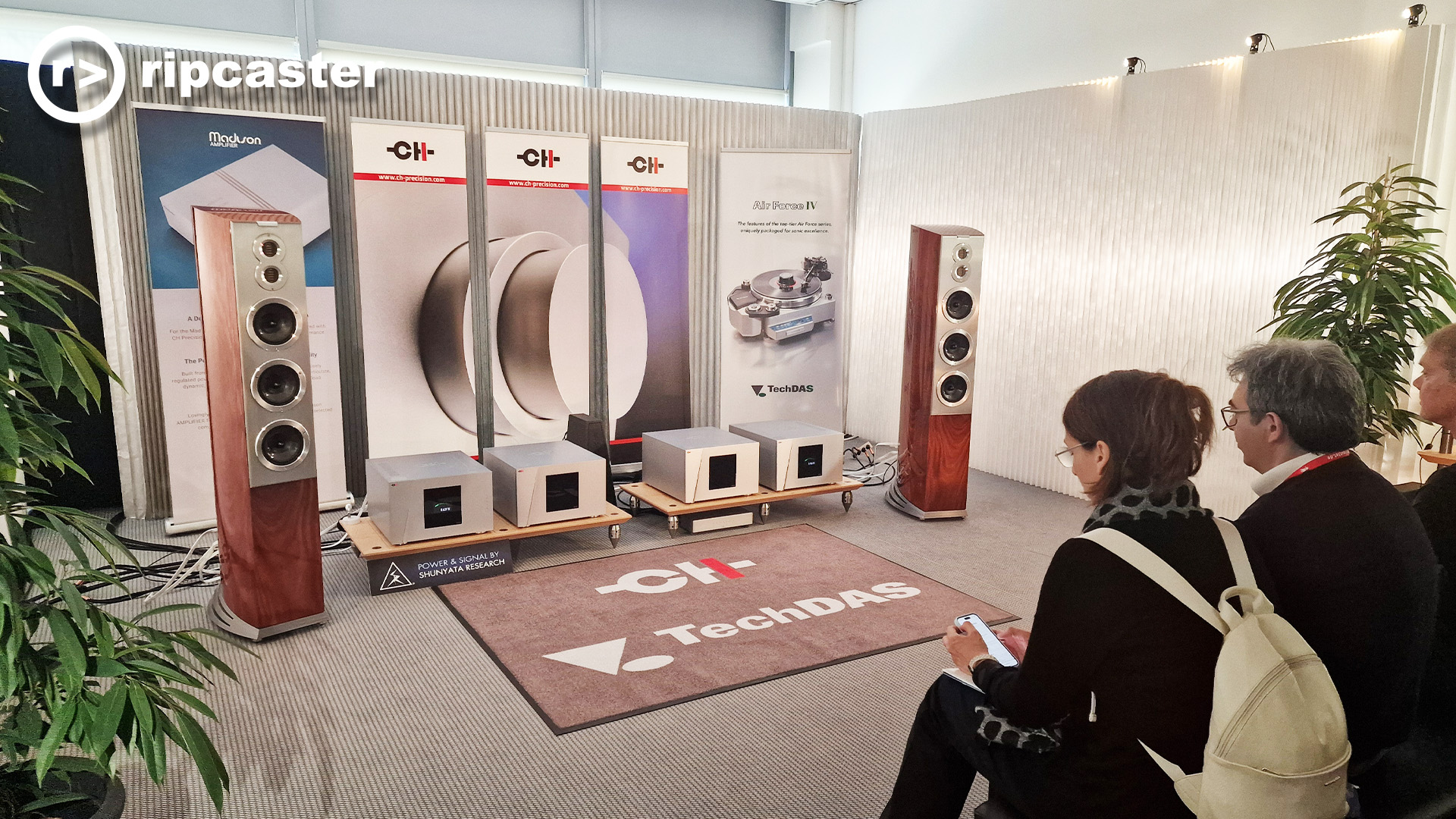 A listening room at the Munich HiFi Show with three people sitting on chairs facing a HiFi set-up with floorstanding speakers either side of various HiFi units.