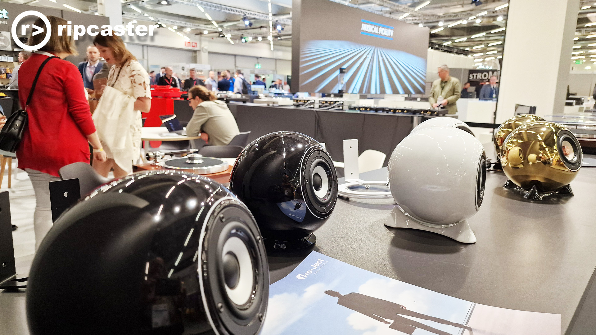 A HiFi stand showing sphere shaped speakers.  Two of them are shiny gold.  There are women in this photo.
