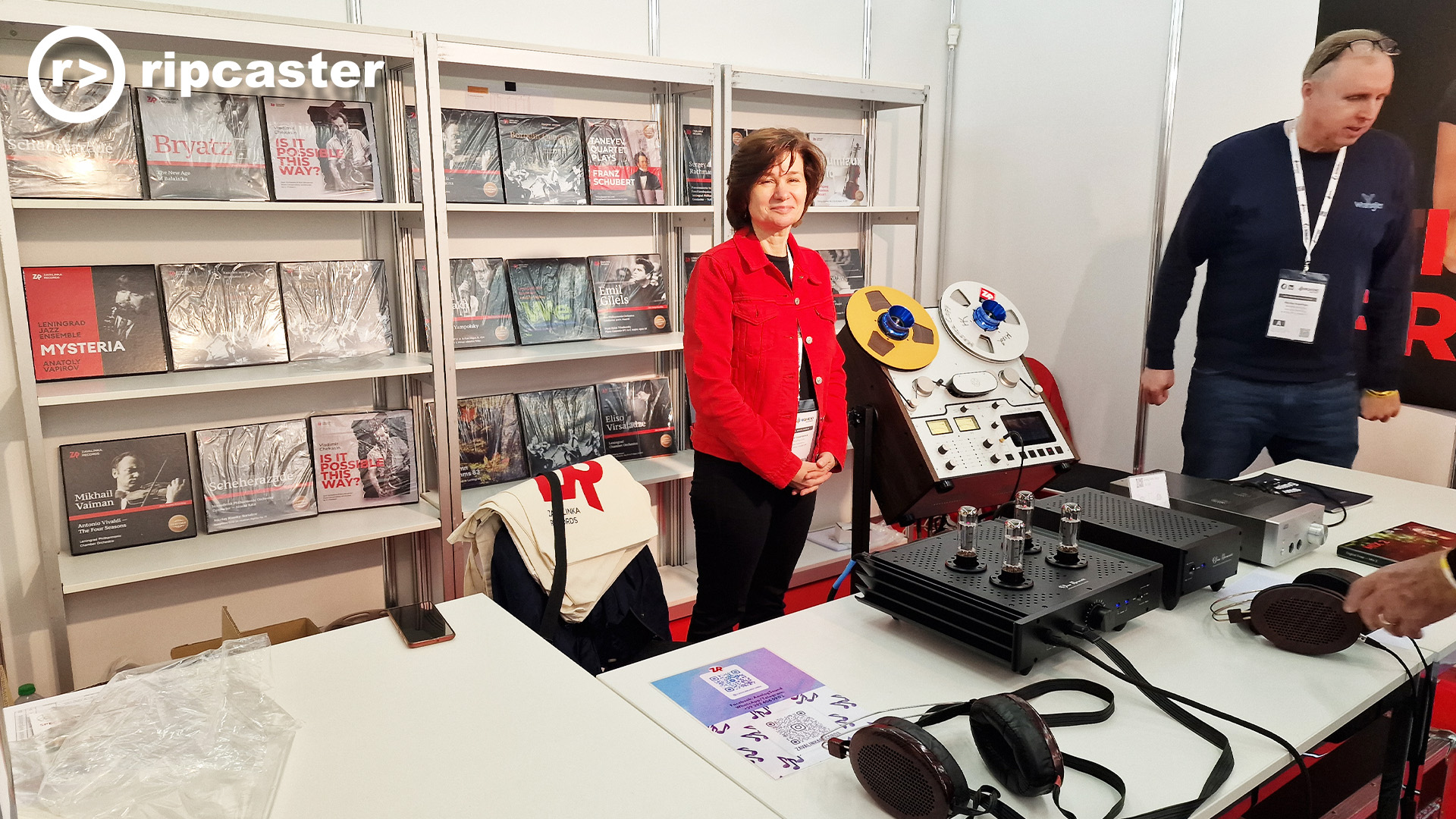 A woman in a red jacket looking at the camera and a man.  they are standing on a reel to reel tape selling stand at the HiFi Munich show.