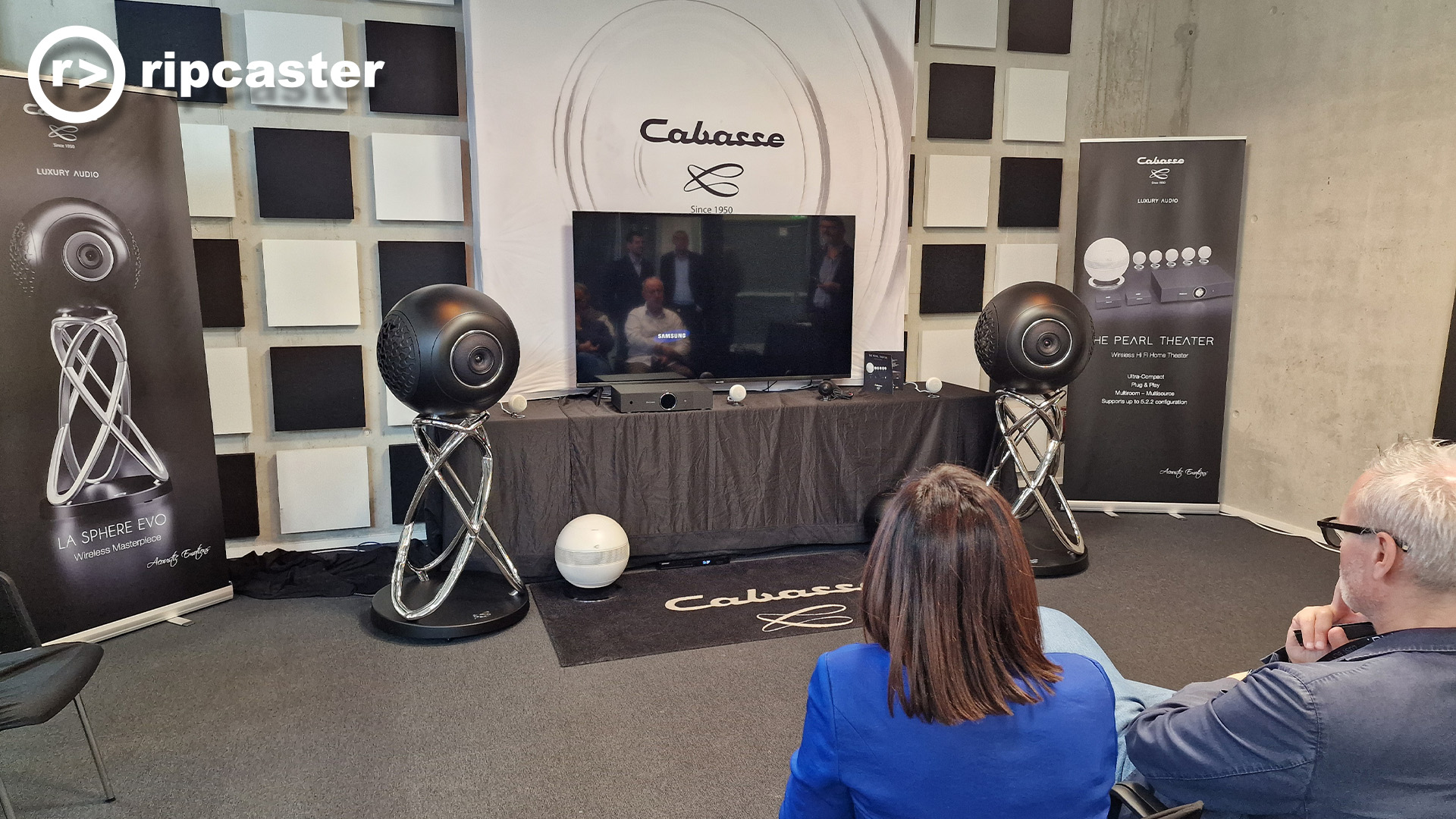The Cabasse room at the HiFi show.  Two people are in the foreground with their backs to us.  there's speakers at the front and other HiFi Units.