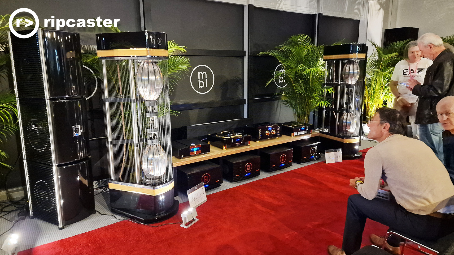 An interesting thing that looks a bit like a stick with a balloon on each end.  The balloon shape on each end is either a light or it's reflecting light.  There are various black HiFi units and men sitting to the right of the image looking towards the audio equipment.