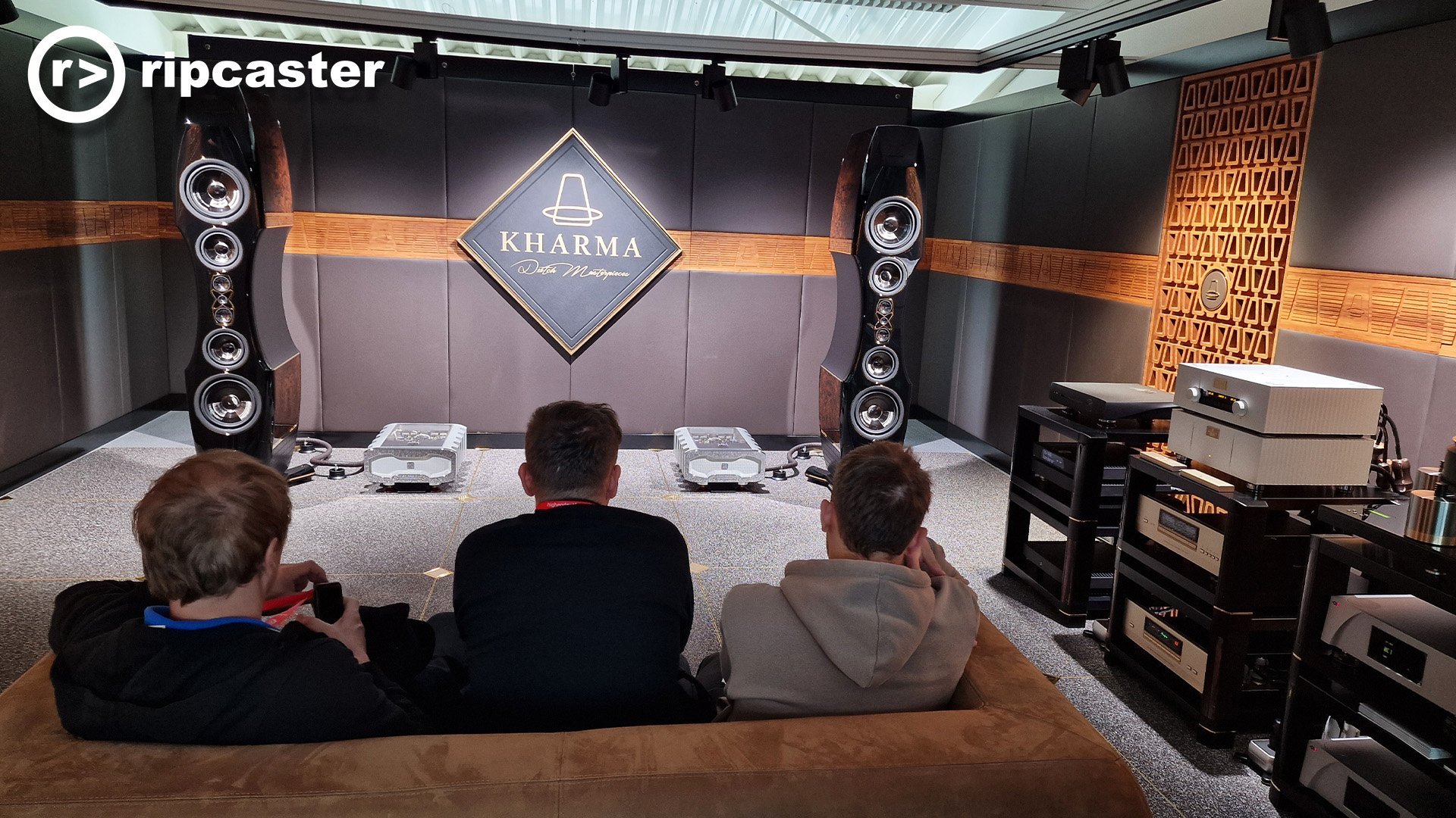 Two large black floorstanding speakers in a demo room at a HiFi show with other equipment around the room.  there are three men viewed from the back on a sofa in the foreground.