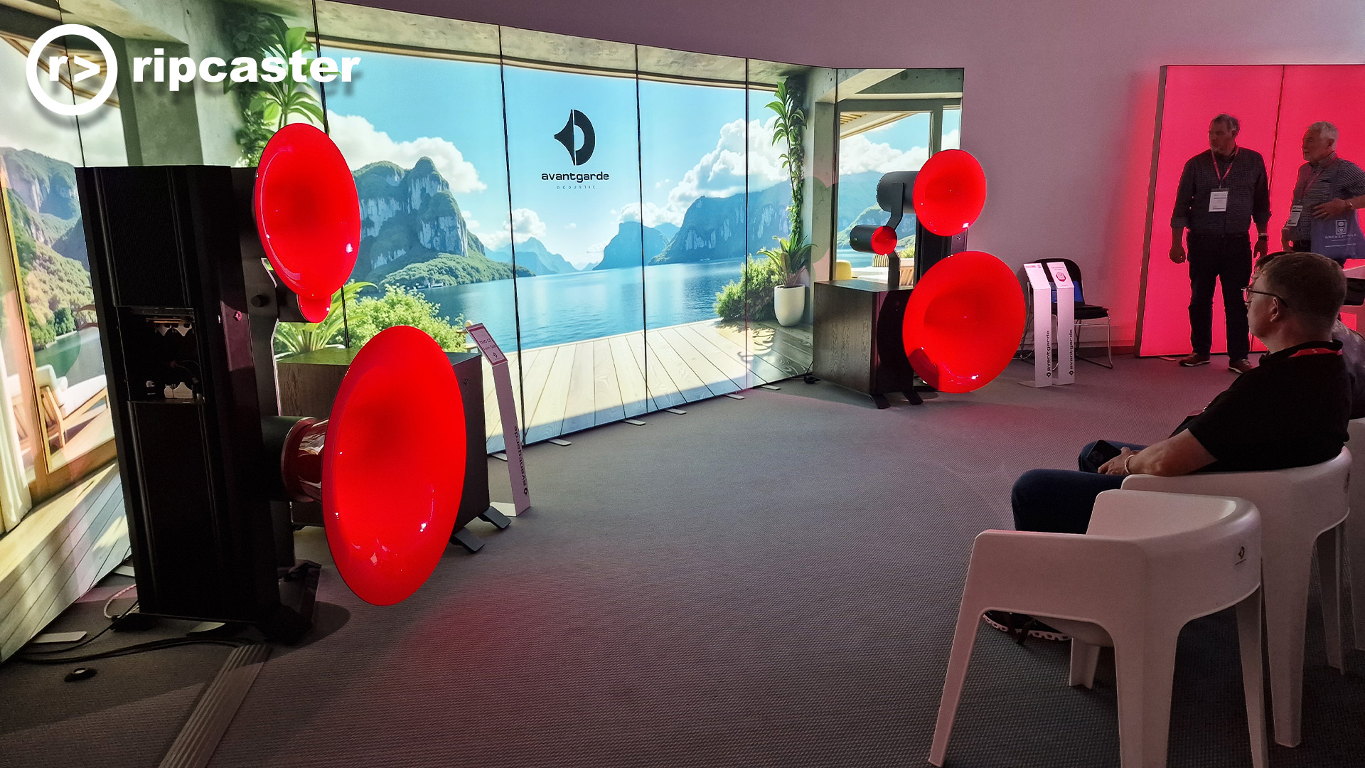 Floorstanding speakers with bright red horns at the Munich HiFi show.  there are men to the right of the image.  Some standing, some sitting.  If I should happen to spot a woman among the images I'm seeing, I will delight in telling you in my description.