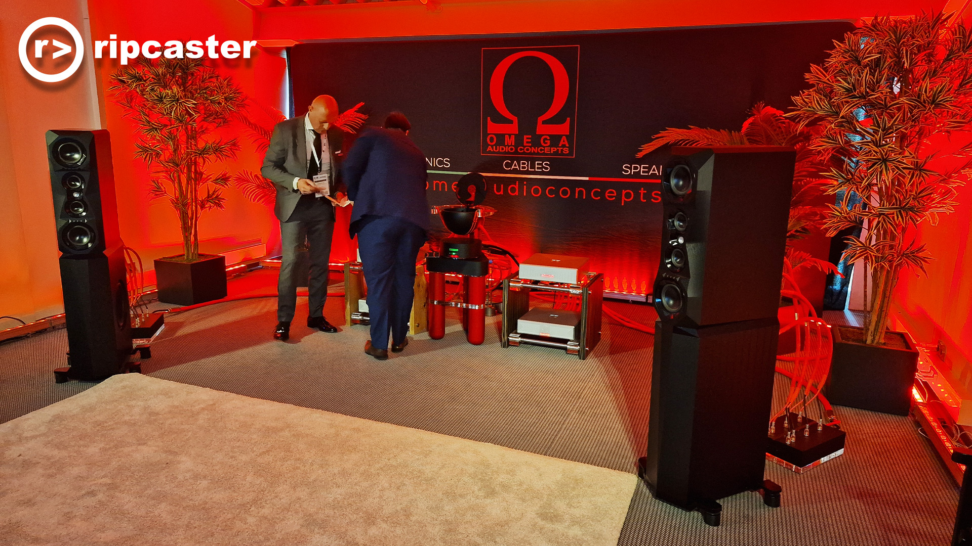 Very red lighting in this image.  Two men stand by HiFi equipment with two black floorstanding speakers either side and some tall plants