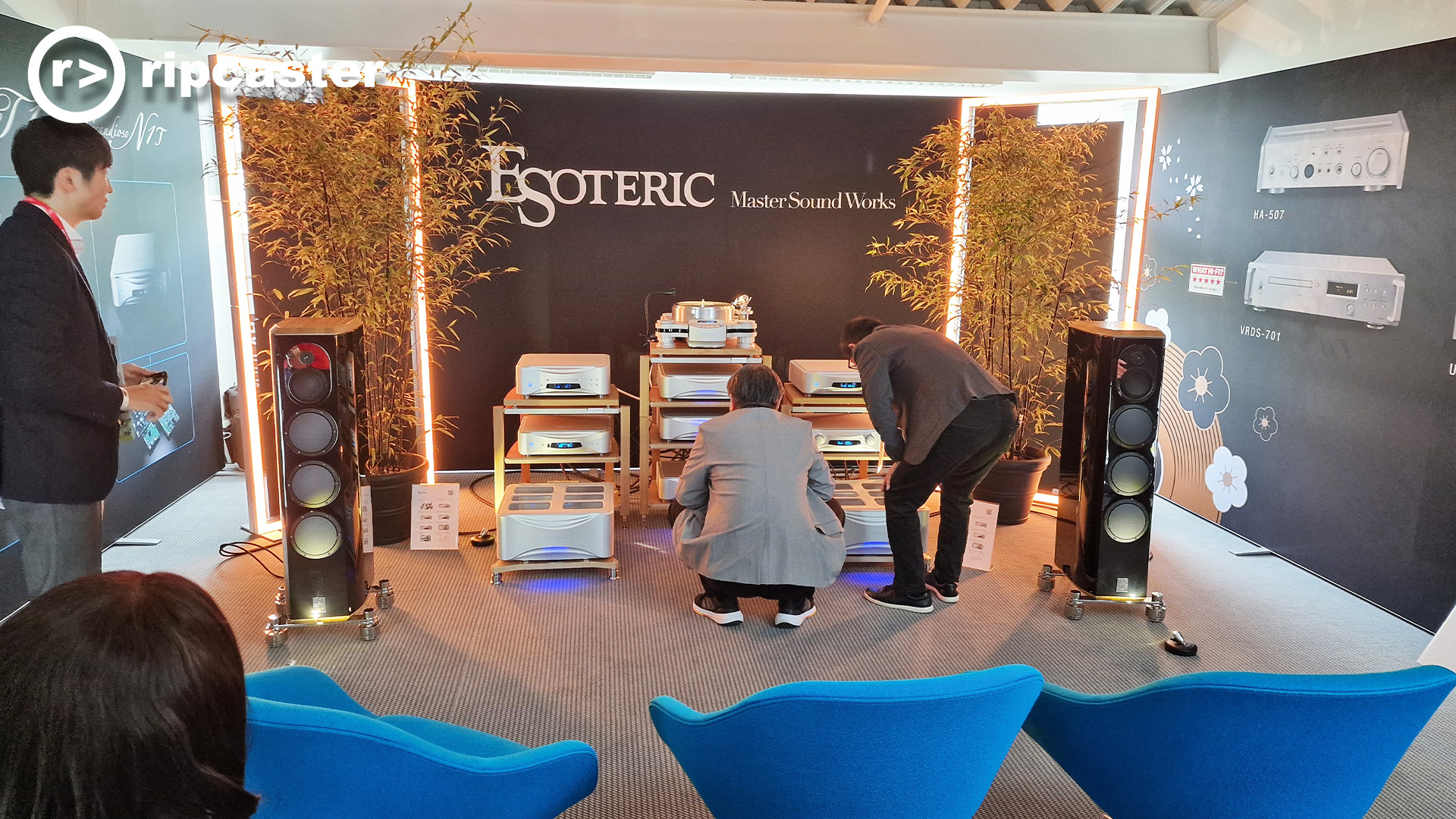 Esoteric.  Two men crouch looking at an array of HiFi units that sit on racks between two black floorstanding speakers.
