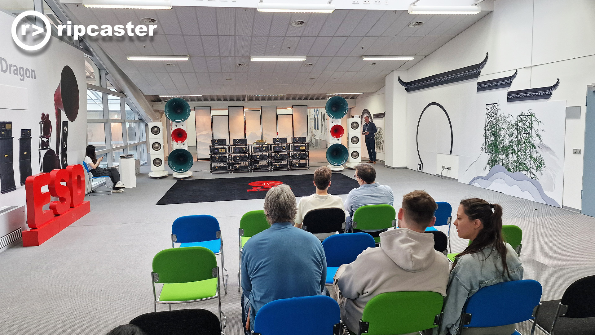 People viewed from the back including two women.  they are sitting on chairs for a demonstration of some speakers with large horns in a large room with a lot of HiFi units 