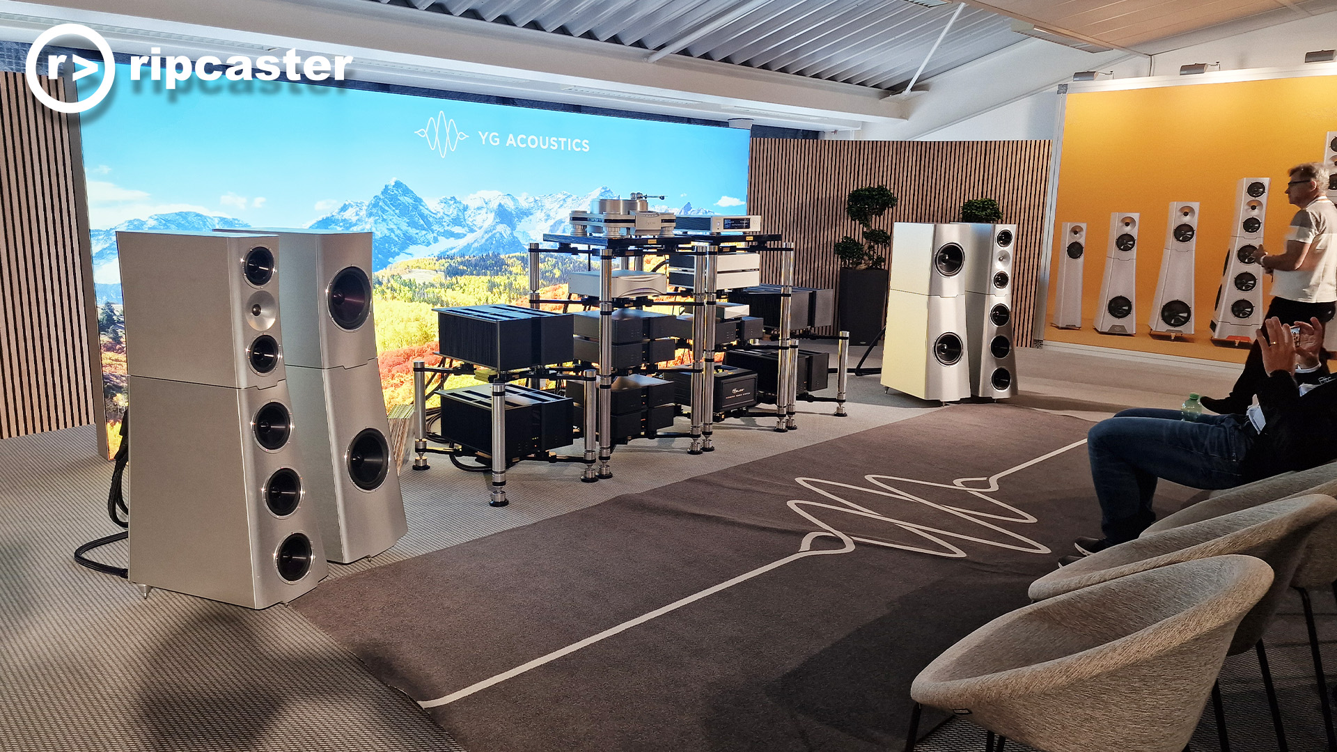 Y G Acoustics.  Large silver floorstanding speakers with a mass of HiFi equipment on HiFi racks in between them and chairs to the right of the image.  Two men can be partially seen on the right of the image.  One sitting taking a photo and one walking towards the speakers