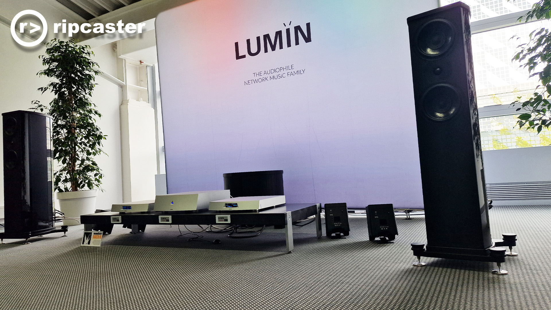The Lumin room at the Munich HiFi show with silver Lumin boxes between a pair of black floorstanding speakers.