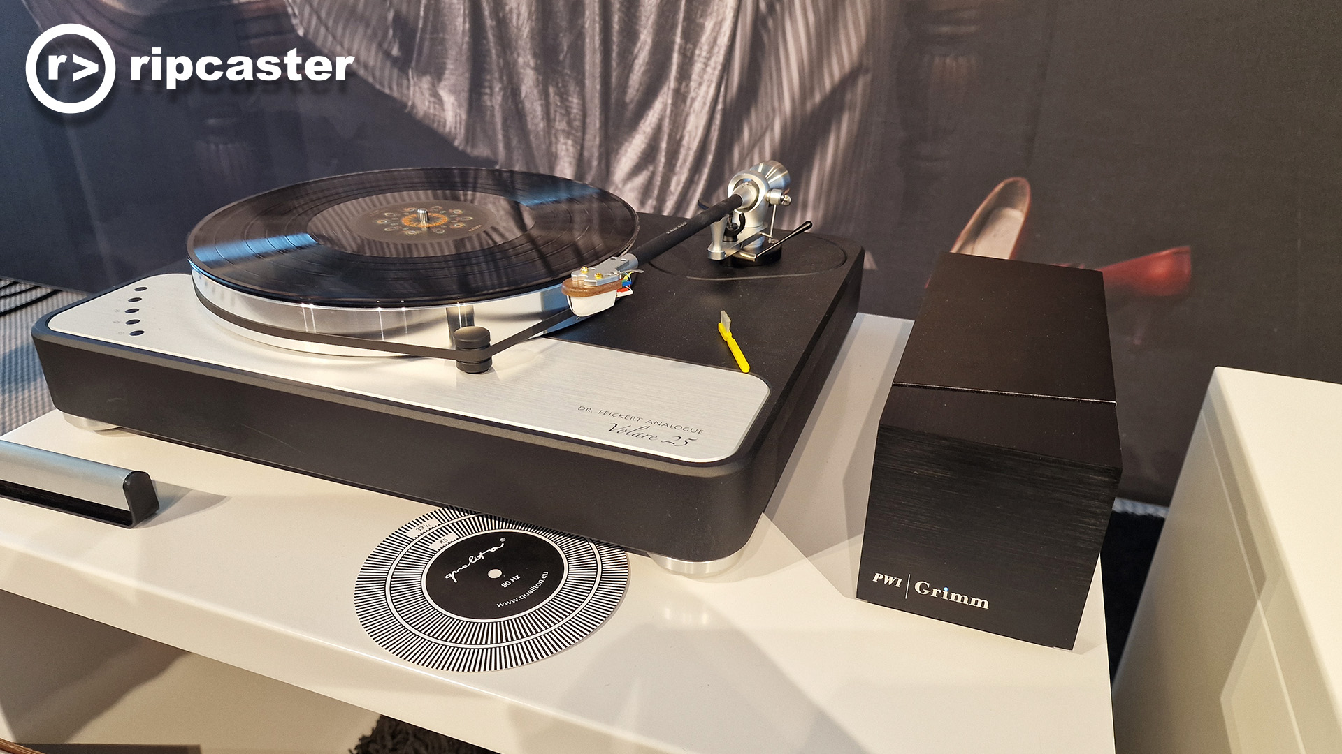 A record player beside a Grimm PW1 on top of a low unit with a backdrop of a woman lounging on a chair