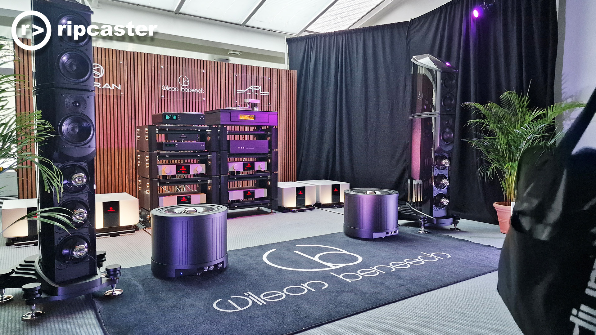 Very large black floorstanding speakers with HiFi kit slightly behind and between them.  In a demo room at HiFi Munich 2025