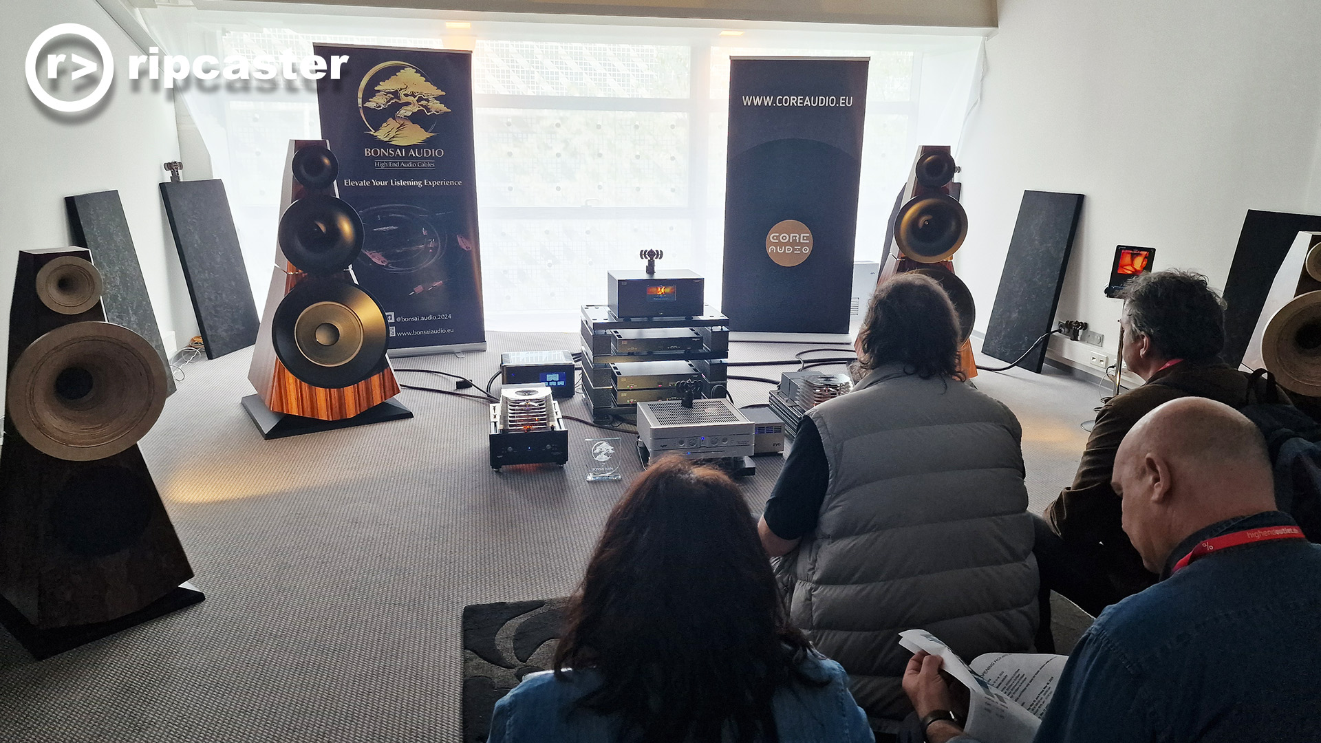 People viewed from behind sitting to listen to a demo in a room at the HiFi show in Munich.  there's a woman which is exciting.  the speakers in this room are wedge shaped: wider at the bottom, than the top and they have dish type bits on the front.