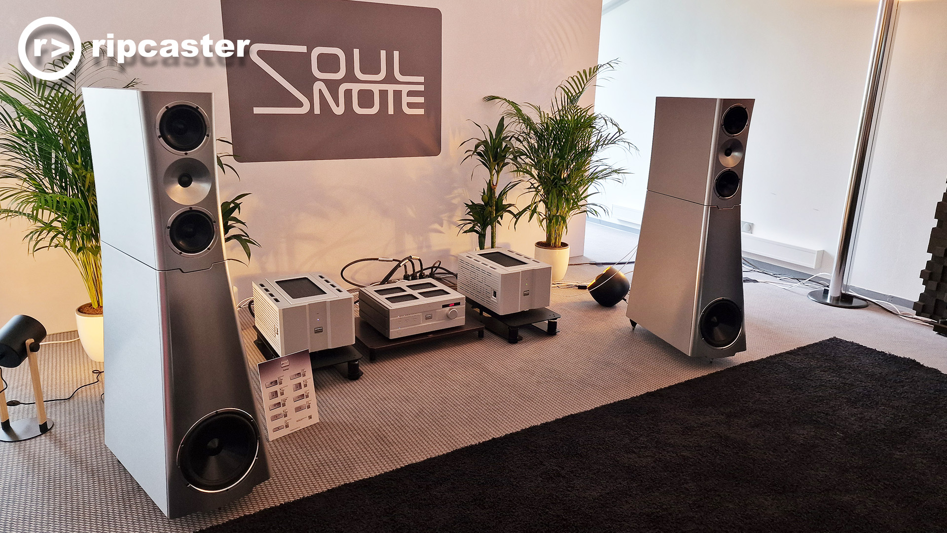 Silver floor standing speakers with silver HiFi kit on the floor between them.  All in a demo room at the HiFi show in Munich.