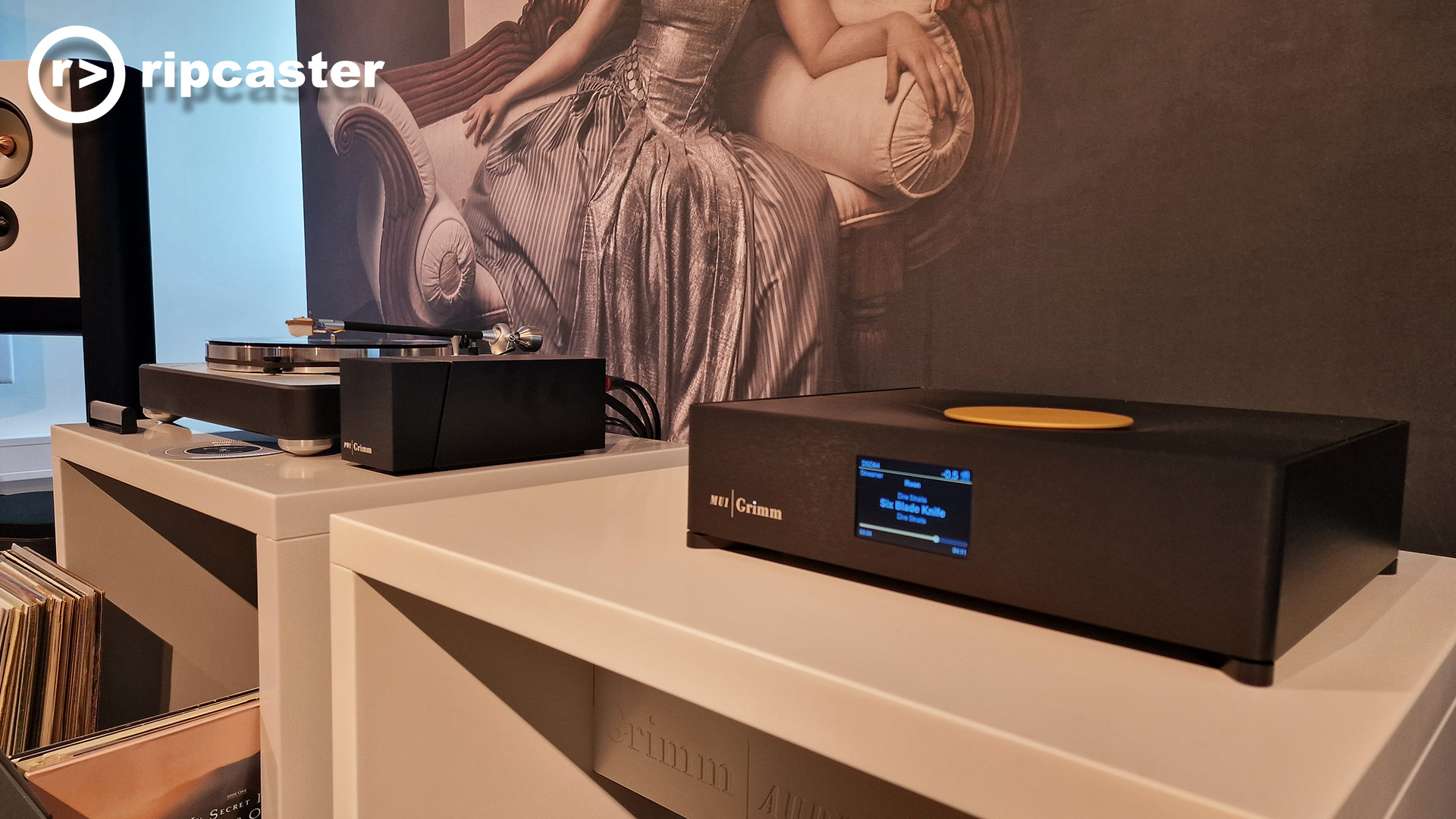 Grimm HiFi equipment on a cream cabinet with a stand mounted speaker to the left and a large backdrop of a woman in a long dress on a chair.