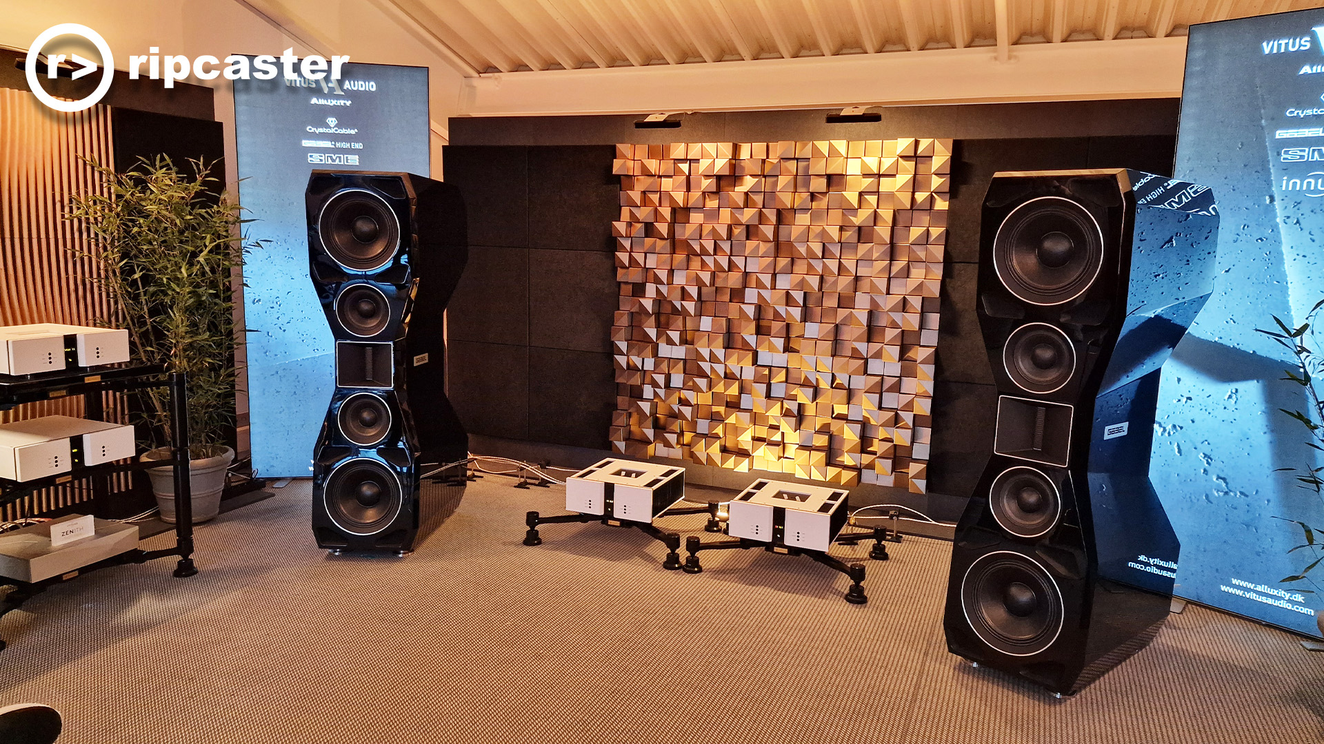 Very large black floorstanding speakers either side of a geometric backdrop with HiFi units between them