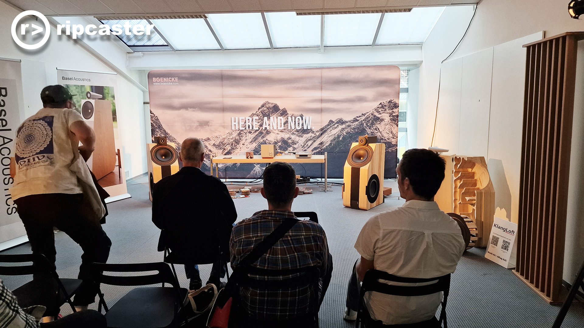 Four men viewed from the back in the same room.  One of them is sitting down or getting up.