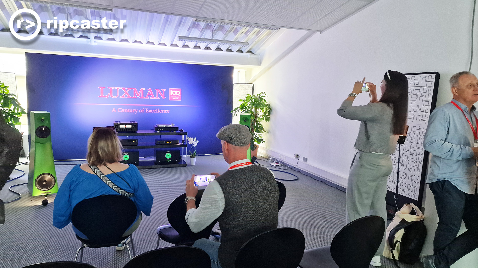 A HiFi demo room at the Munich audio show.  there are two men and two women in this photo. The floorstanding speakers are green.