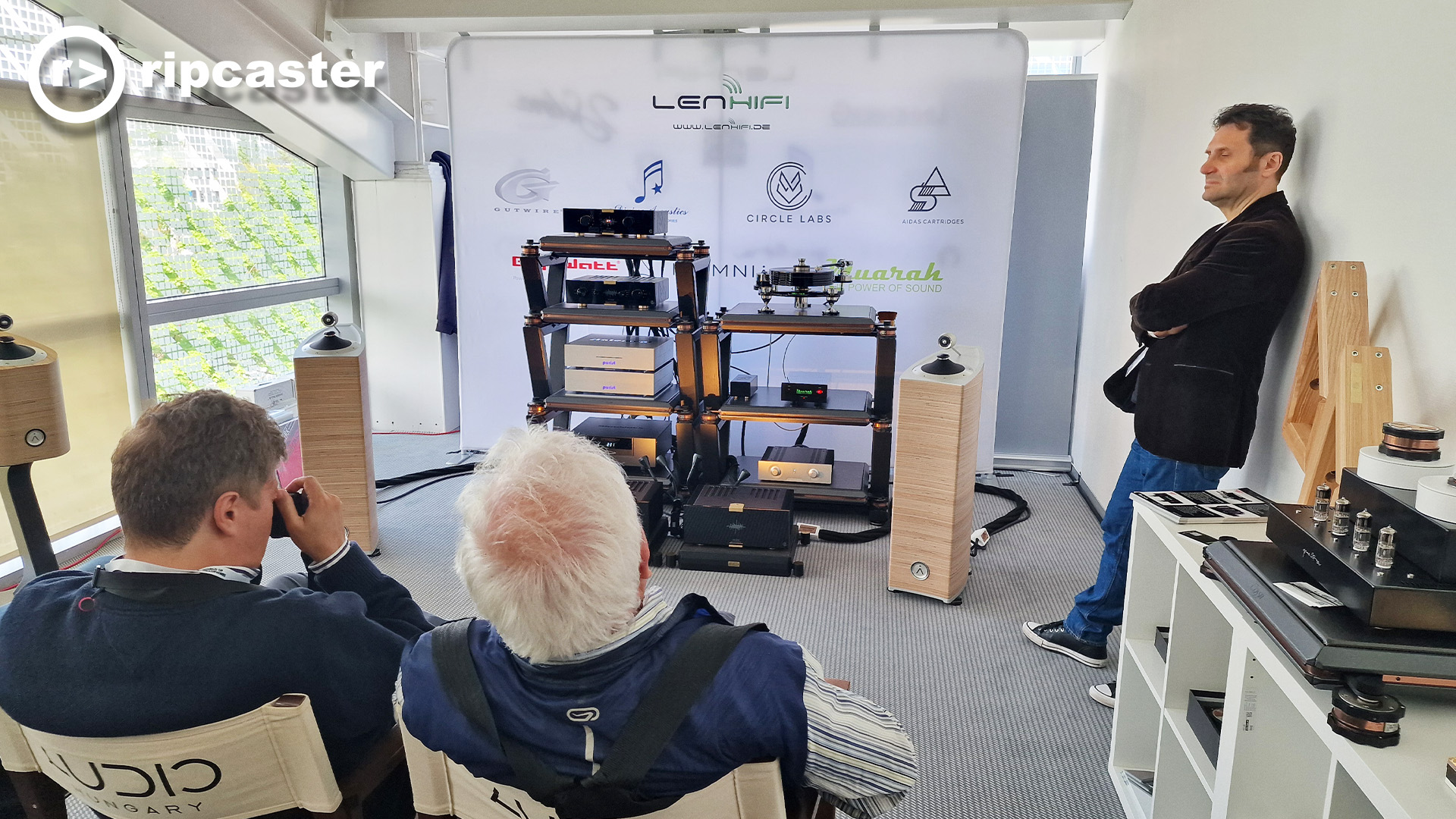 Two men viewed from behind in this listening room and one man standing leaning against the wall.  There's HiFi kit between floorstanding speakers
