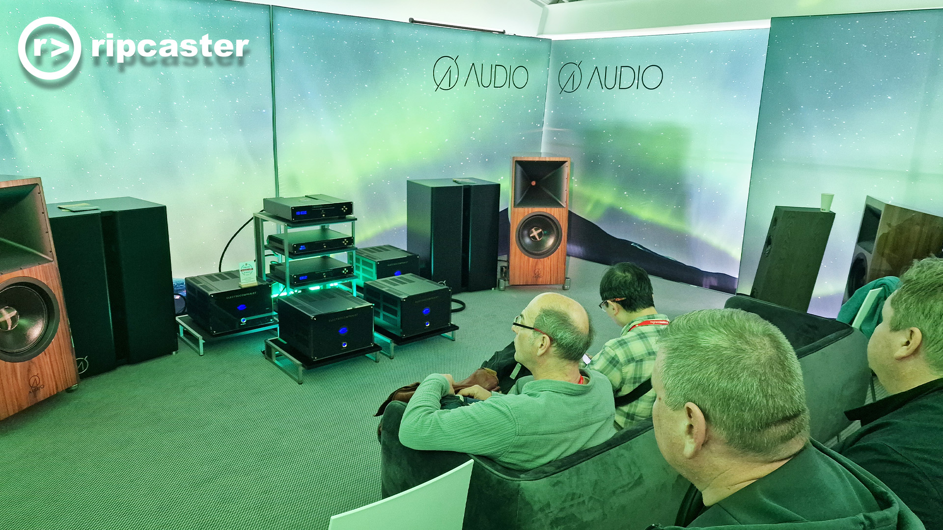 This whole photo has a green tint to it which must be the lighting.  There are four men viewed from the back looking towards a lot of HiFi equipment at the front of the room