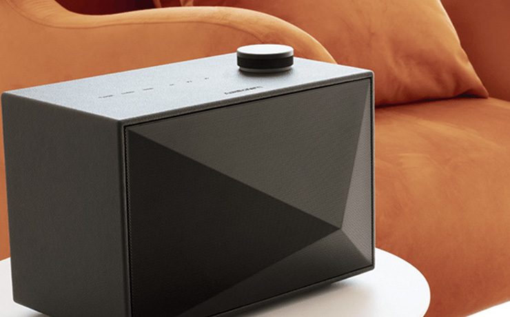 A black rectangular speaker on a white table with an orange sofa behind it.