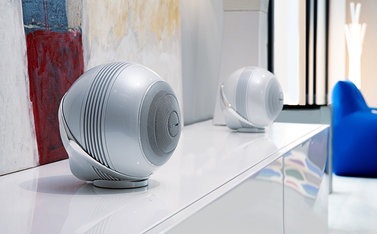 A pair of Cabasse Pearl Akoya speakers in white on top of a white side unit.  The speakers are spherical in shape.  There's a blue chair in the background