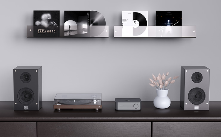 The WiiM Amp Pro on a side unit beside a turntable and with a bookshelf speaker each side. On the wall behind are shelves with albums on them.