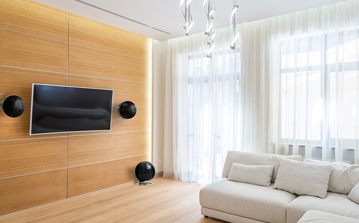 Cabasse Baltic 5 on-wall speakers either side of a tv mounted on a wooden wall and a sub on the floor. A cream coloured corner sofa on the right