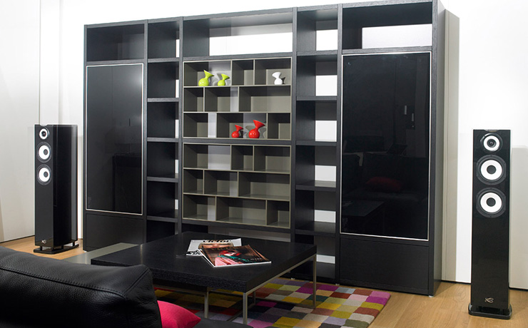 A pair of Cabasse Java MC40 floorstanding speakers either side of a large shelving unit.