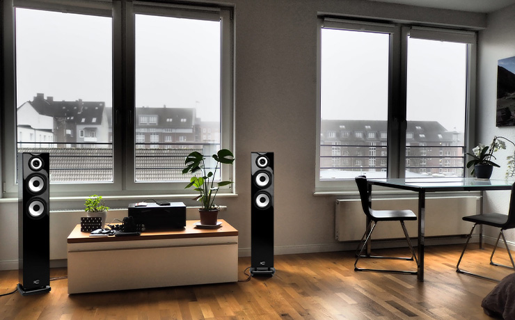 A pair of Cabasse Java MC40 floorstanding speakers in black either side of a low unit that has houseplants and a black unit on top.  There are large windows looking out onto groups of flats.  There's a table and chairs to the right of the image.