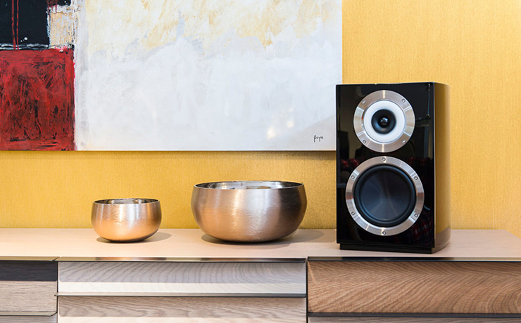 The Cabasse Murano bookshelf speaker in black on a side unit with metal bowls beside it