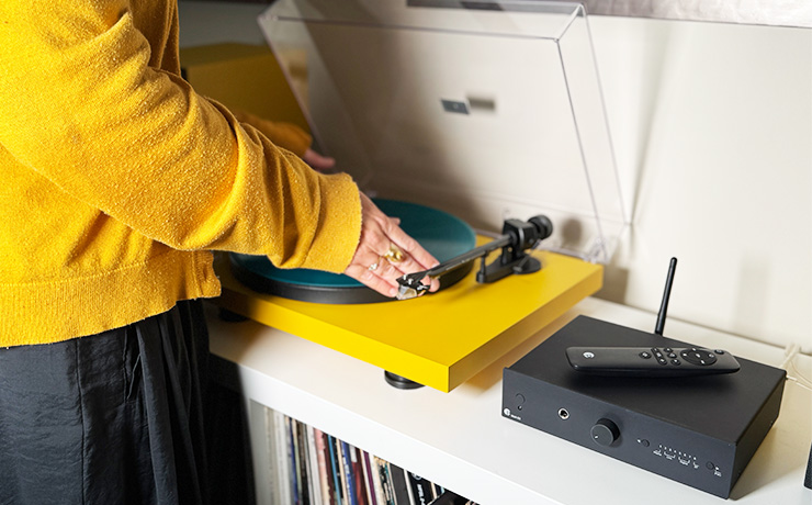 The yellow Project turntable with the Amplifier beside it. A woman in a yellow jumper is putting a green record on the turntable. This is our colourful audio system at the ripcaster showroom
