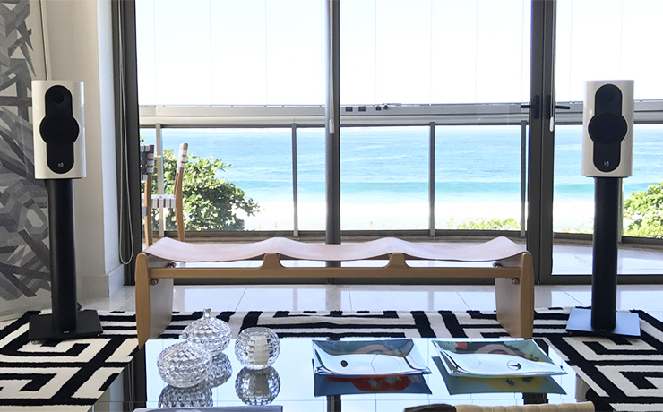 A pair of Kii Three Speakers in white in front of a large window that looks out to a beach with a very clear looking blue sea. In the foreground there's a coffee table with various glass items on it. The rug is a geometric black and white pattern and there's a bench with a canvas seat