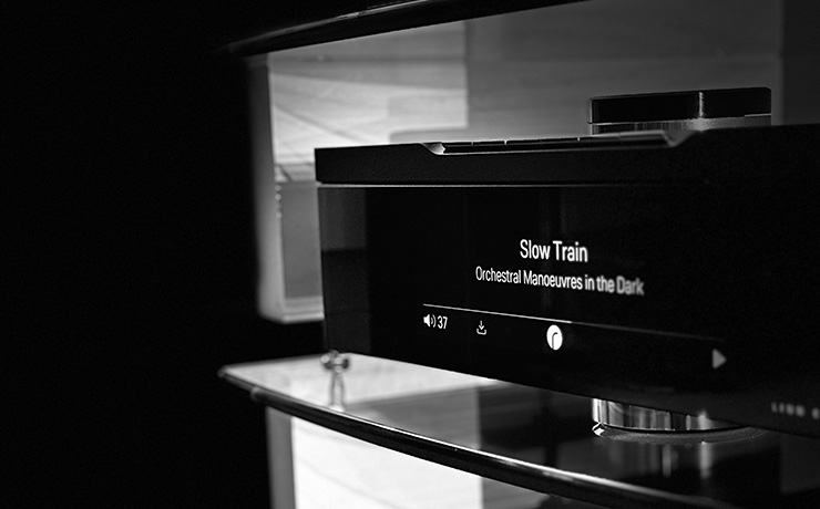 The Linn Klimax DSM on a glass shelf showing the track playing on the screen which is 'Slow Train' by OMD. The image is black and white