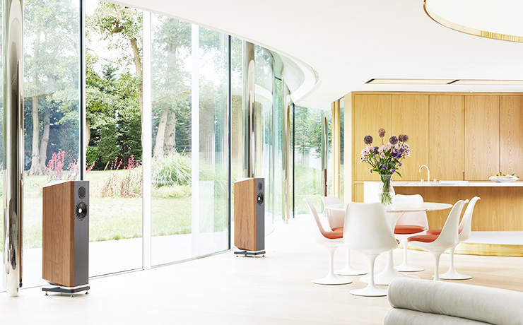 Two large floor standing speakers in front of very large glass windows. There's a round table with white modern chairs around it.