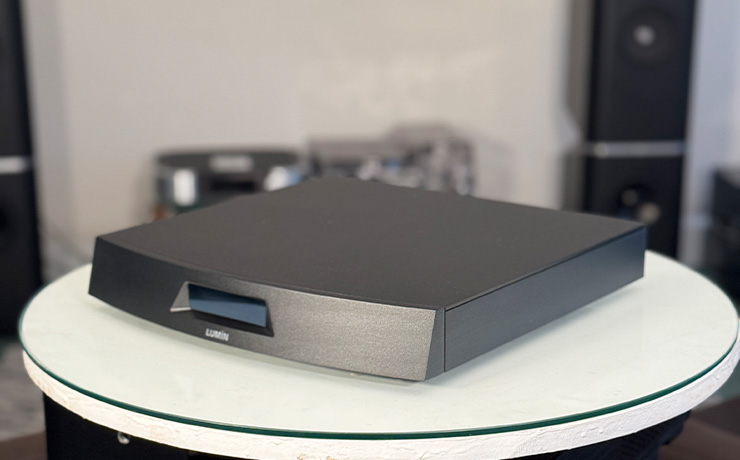 The Lumin L2 in black at the ripcaster showroom.  It's on a white circular stand with a glass top.  Behind it are various pieces of HiFi equipment including the Kudos 808 floorstanding speakers.