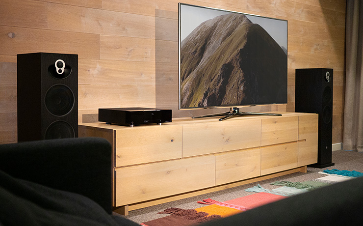 A pair of black Majik 140 speakers either side of a low level unit with a tv and a DSM on it.