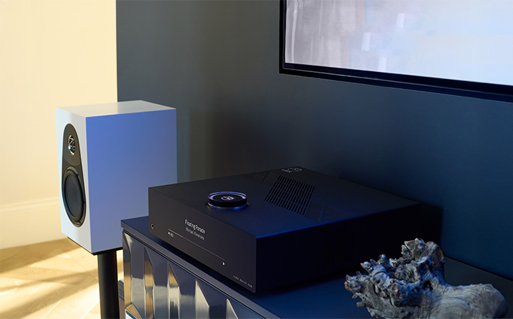 Linn Majik DSM - 5th Generation (2025) on a side unit with a white Linn 119 speaker beside it on a stand. The wall behind the DSM is very dark blue with a picture on the wall that's only partly visible. Beside the DSM is what looks like a lump of driftwood. There's light streaming in to the rear left of the image. the light falls onto a wooden floor and a light coloured wall