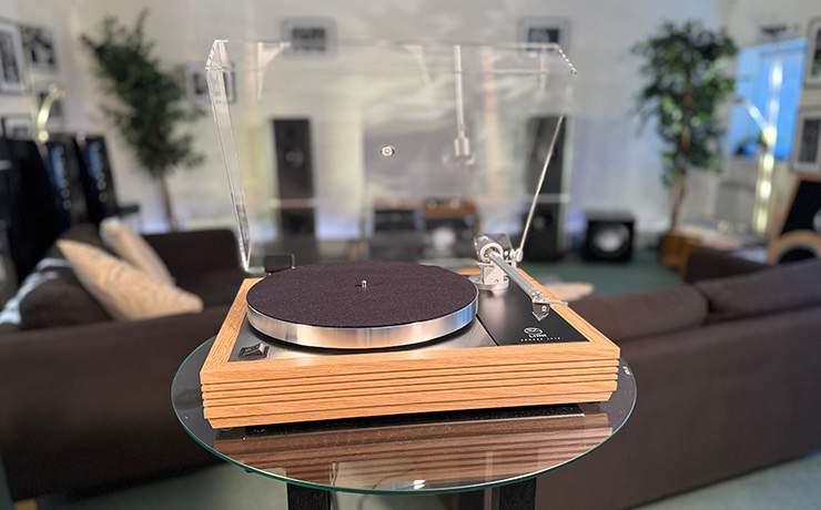 The Linn Majik LP12 MM Turntable with a fluted plinth in oak. The lid is raised. Behind the turntable are speakers and sofas along with other HiFi equipment and some large indoor plants