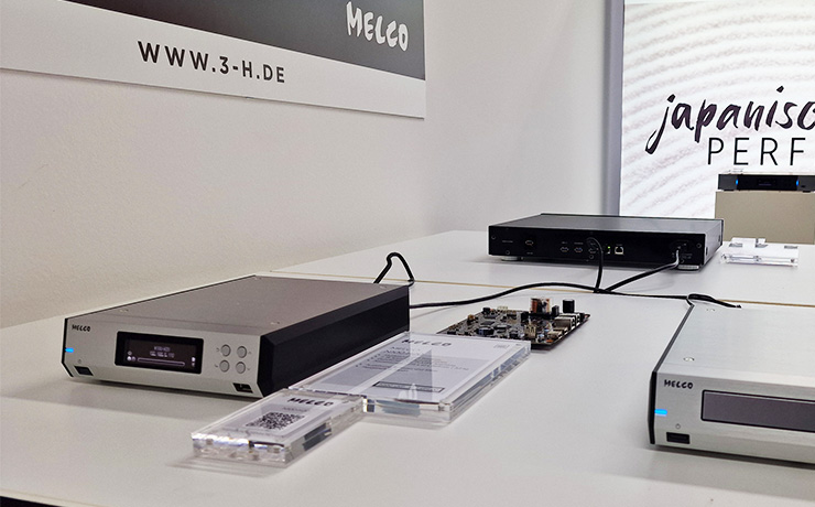 The Melco N100 on a white table at the Munich 2024 HiFi show beside another Melco box on a white table. The N100 is a silver box that looks the shape of an old answer machine. It has a screen at the front and four buttons to the right.