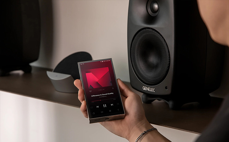 Astell & Kern PD10 in someone's hand with the cradle on a shelf and a bookshelf speaker beside it