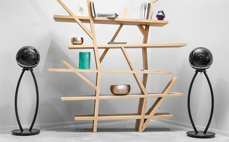 A pair of Cabasse Pearl loudspeakers in black on stands either side of a wooden shelving unit