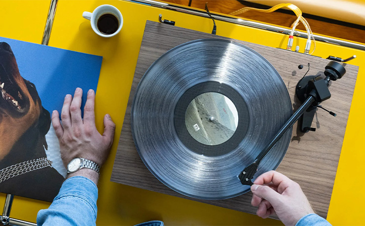 The Project EVO 2 turntable in walnut on a yellow table with a man's hand putting the needle on a record