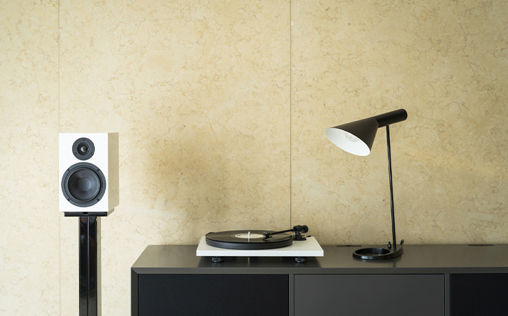 The Project Primary E turntable in white on a black unit with a lamp one side and a bookshelf speaker on a stand the other side.
