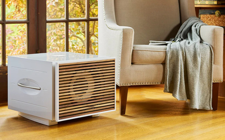 A white REL S/550 subwoofer in a living space.  It has a wooden grille and is beside a beige armchair