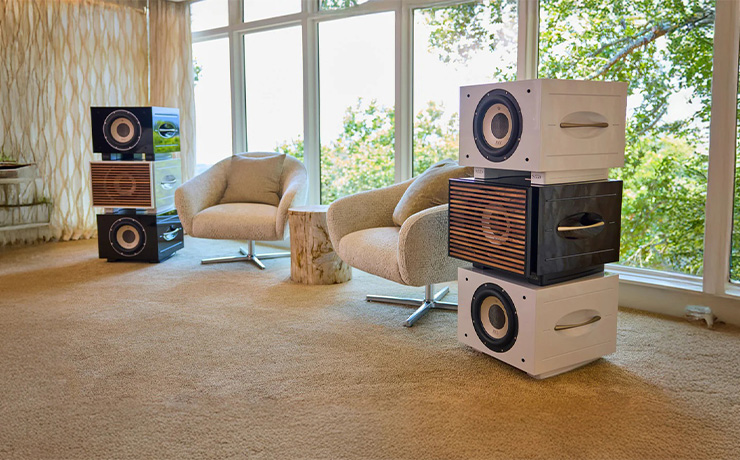 Two stacks of three REL S/550 subwoofers in front of a wall of windows.  The room is carpeted in beige and there are two swivel armchairs between the stacks.  The left stack has two black woofers with no grille with a white one with a wooden grille in the middle.  On the right is a black one with a wooden grille in the middle with a white on top and a white underneath