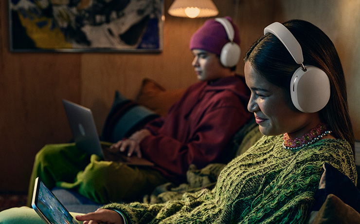 Two people sitting on a sofa wearing white Sonos Ace headphones and both looking at their individual screens