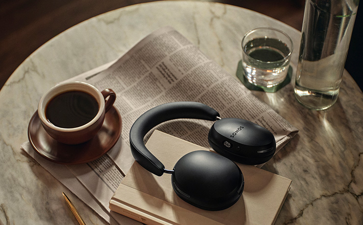 A black pair of Sonos ACE headphones laying on a table that has a cup of coffee, water, a newspaper, book and pen