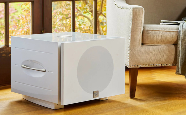 A white REL S/850 on a wooden floor in a living space. The grille is cloth